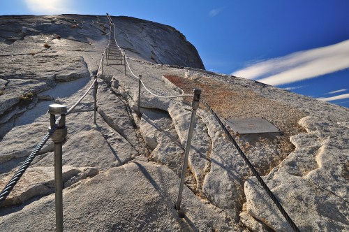 A steep granite slope with metal cables for climbing, under a clear blue sky with wispy clouds.