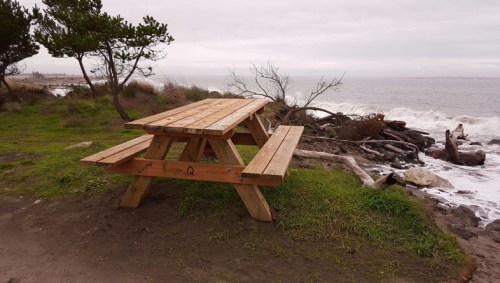 A wooden picnic table by the shore, with waves crashing against rocks and a cloudy sky in the background.