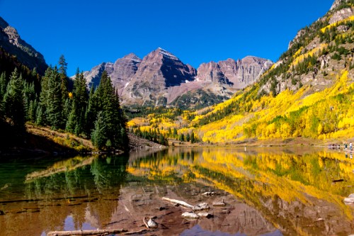 A serene lake reflects vibrant autumn foliage and towering mountains under a clear blue sky.