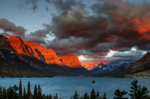 Sunset over a lake surrounded by mountains, with dramatic clouds and vibrant orange and red hues illuminating the peaks.