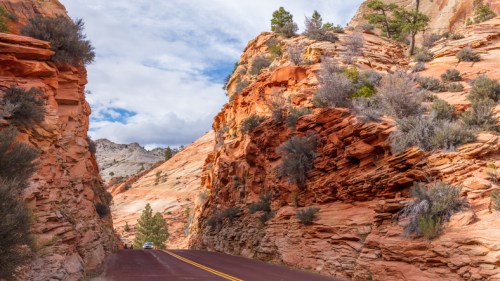 A winding road cuts through vibrant red rock formations under a partly cloudy sky. Lush greenery peeks from the cliffs.
