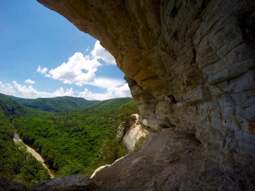 A scenic view from a rocky cliff overlooking lush green hills and a winding river under a bright blue sky.