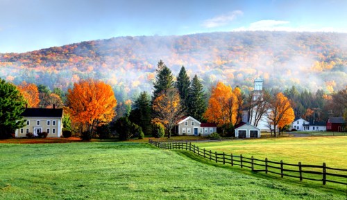A picturesque autumn landscape with colorful trees, a misty mountain, and charming houses beside a green field.