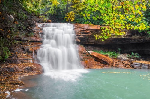 A serene waterfall cascades over rocks into a tranquil pool, surrounded by vibrant autumn foliage.