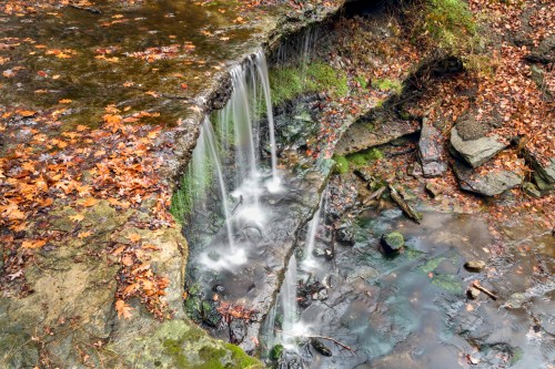 A serene waterfall cascading over rocks, surrounded by autumn leaves and a tranquil stream below.
