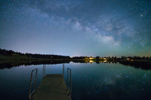 A serene lake at night reflecting a starry sky and the Milky Way, with a dock extending into the water.