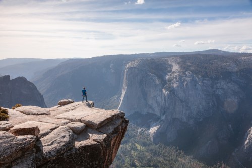 A person stands on a rocky ledge overlooking a vast mountain landscape under a clear blue sky.