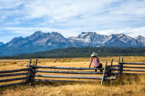 A person in a hat sits on a wooden fence, overlooking a vast meadow with mountains in the background under a blue sky.