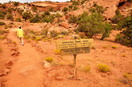 A hiker walks past a trail sign for Upheaval Dome and Syncline Loop in a rocky, desert landscape.