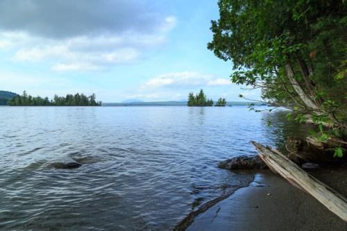 A serene lakeside view with calm water, distant islands, and trees lining the shore under a partly cloudy sky.