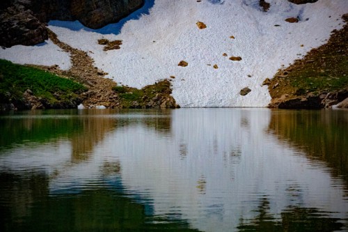 A serene lake reflecting snow-capped mountains and green hills under a clear sky.