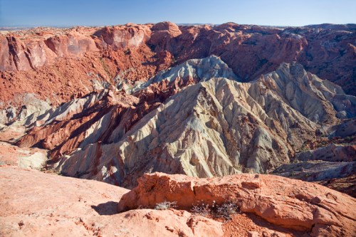 A panoramic view of colorful, layered rock formations in a rugged canyon landscape under a clear blue sky.