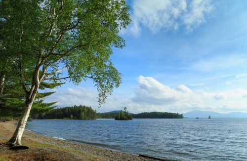 A serene lakeside view with trees, a sandy shore, and distant islands under a partly cloudy sky.
