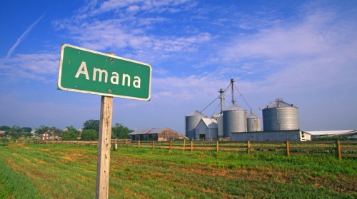 A green sign reading "Amana" in front of silos and a barn under a blue sky with scattered clouds.