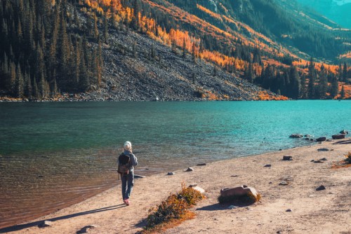 A person walks along a sandy shore by a vibrant lake surrounded by autumn-colored trees and mountains.