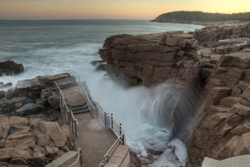 A rocky coastline with waves crashing against cliffs at sunset, featuring a pathway leading to the water.
