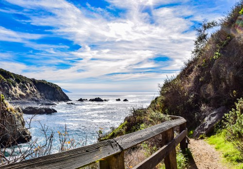A scenic coastal view with a wooden railing, rocky shoreline, and bright blue sky with wispy clouds.