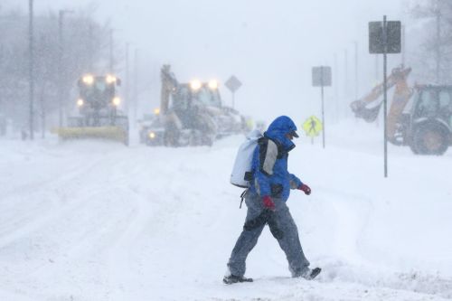 A person walks through deep snow while snowplows clear the road in a winter storm. Visibility is low due to heavy snowfall.