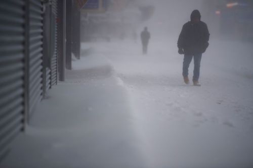A person walks through a snowy street during a blizzard, with another figure faintly visible in the background.