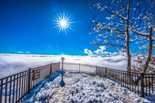 A snowy overlook with a bright sun shining above clouds, surrounded by a railing and a tree with snow-covered branches.