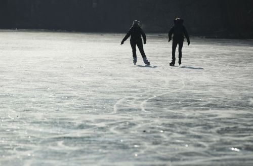 Two people ice skating on a frozen lake, with sunlight casting long shadows on the icy surface.