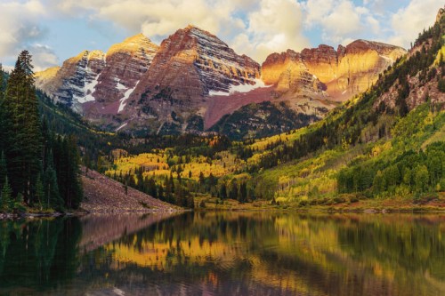 Scenic view of mountains reflecting in a calm lake, surrounded by vibrant autumn foliage and a clear sky.