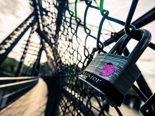 A close-up of a padlock on a chain-link fence, with a blurred bridge in the background under a cloudy sky.