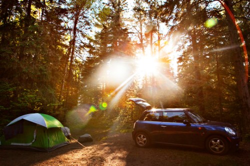 A blue car parked near a green tent in a sunlit forest, with sunlight streaming through the trees.