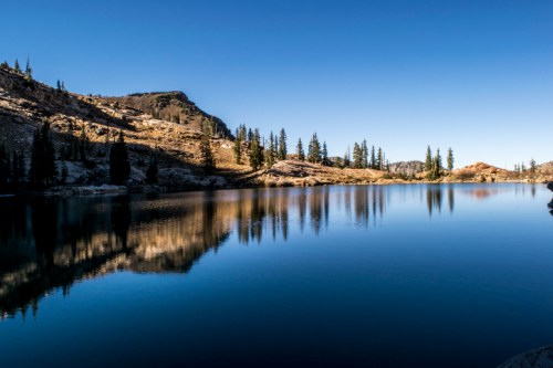 A serene lake reflecting surrounding mountains and trees under a clear blue sky.