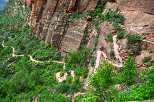 Winding hiking trail along a steep, rocky cliff surrounded by lush greenery in a mountainous landscape.
