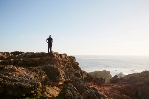 A person stands on rocky terrain, gazing out at the ocean under a clear sky.