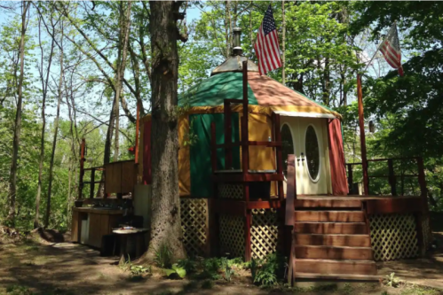 A colorful yurt nestled among trees, featuring a wooden deck and an American flag flying above.