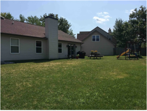 A sunny backyard with a house, picnic table, flower pots, and a playground set on green grass.