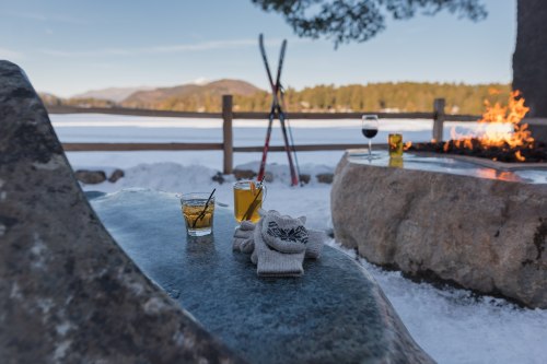 Two drinks on a stone table by a snowy landscape, with skis in the background and a fire pit nearby.