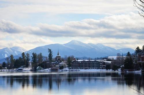 A serene winter landscape featuring a lake, mountains, and a quaint town with snow-covered trees and buildings.