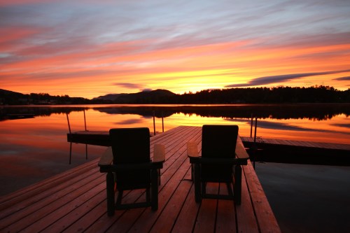 Two wooden chairs on a dock overlooking a tranquil lake at sunset, with vibrant orange and purple skies reflected in the water.