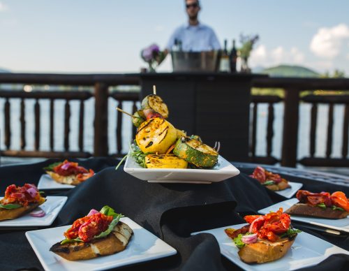 A platter of grilled vegetables and bruschetta on a table, with a blurred bartender and scenic waterfront in the background.