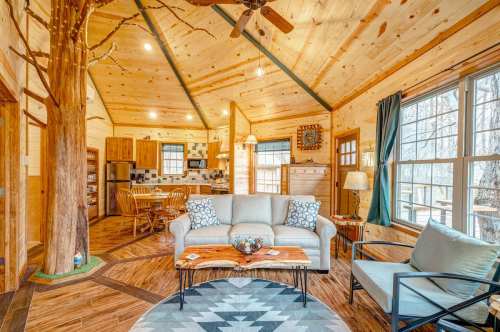 Cozy cabin interior with wooden walls, a tree pillar, comfortable seating, and a dining area visible in the background.