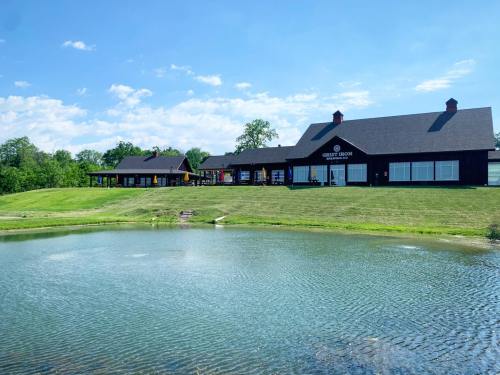 A modern building with large windows sits by a pond, surrounded by green grass and trees under a blue sky.