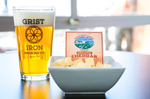 A glass of golden beer next to a bowl of cubed cheddar cheese and a cheese package on a table.