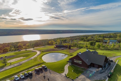 Aerial view of a scenic landscape with a lake, green fields, and a modern building under a cloudy sky at sunset.