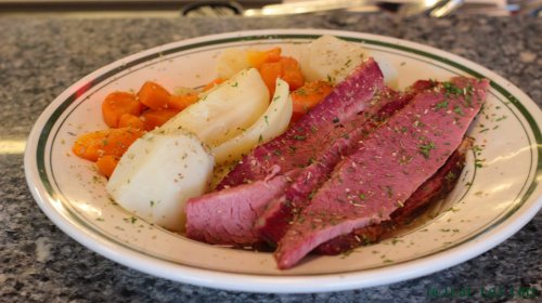 A plate of sliced corned beef with carrots and potatoes, garnished with herbs, on a marble countertop.