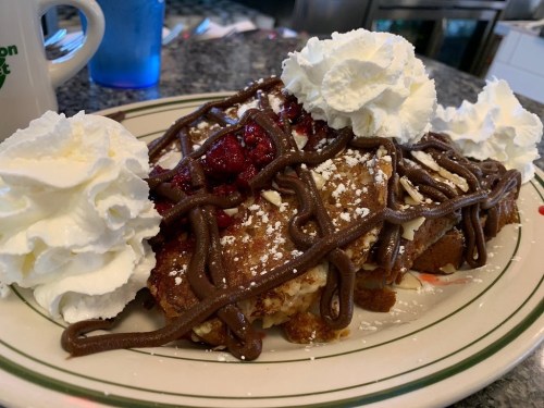 A plate of dessert featuring layers of cake topped with whipped cream, chocolate drizzle, and raspberry sauce.