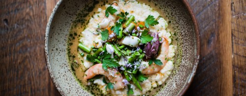 A bowl of creamy shrimp and grits topped with fresh herbs and vegetables, set on a wooden table.