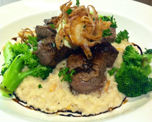 A plate of tender beef medallions on creamy risotto, garnished with crispy onions and fresh broccoli.