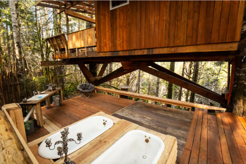 A wooden treehouse with a deck featuring two bathtubs, surrounded by trees in a serene forest setting.
