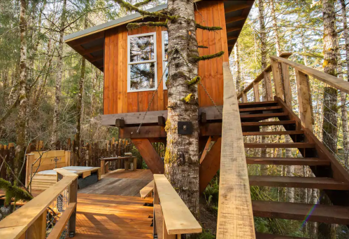 A wooden treehouse nestled among trees, featuring a staircase and outdoor seating area.