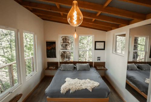 Cozy bedroom with a large bed, wooden beams, and windows overlooking trees, featuring a pendant light and a mirror.