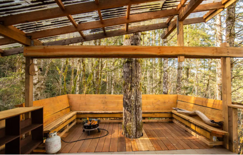Cozy wooden seating area with a central tree, surrounded by forest, featuring a fire pit and rustic decor.