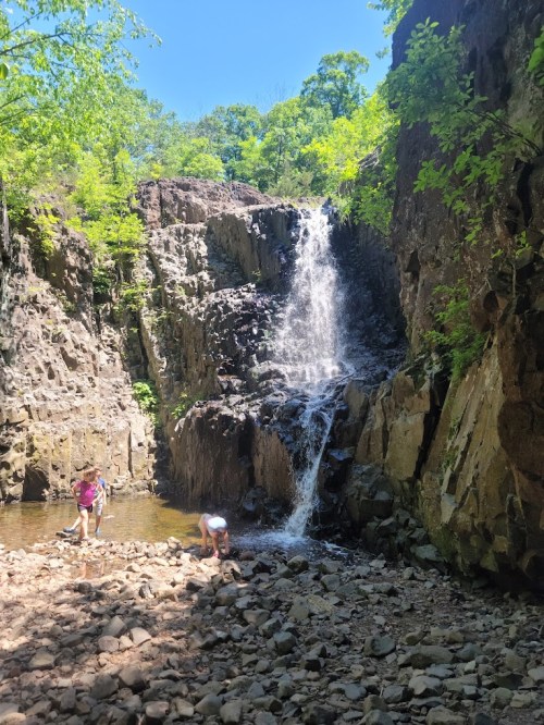 A waterfall cascades down rocky cliffs, with two people exploring the rocky shore and shallow water below.